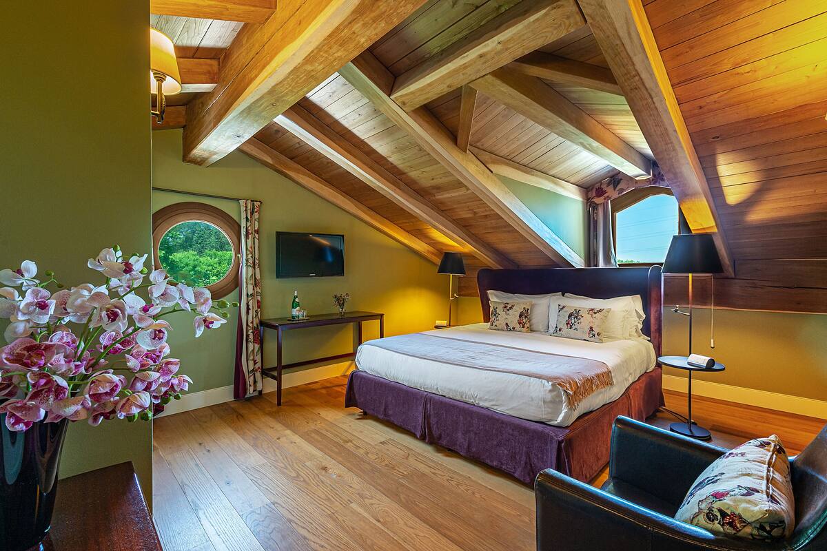 Un dormitorio con techo de madera, una cama con ropa de cama blanca, una mesita de noche, una silla y una ventana con vistas a la vegetación.