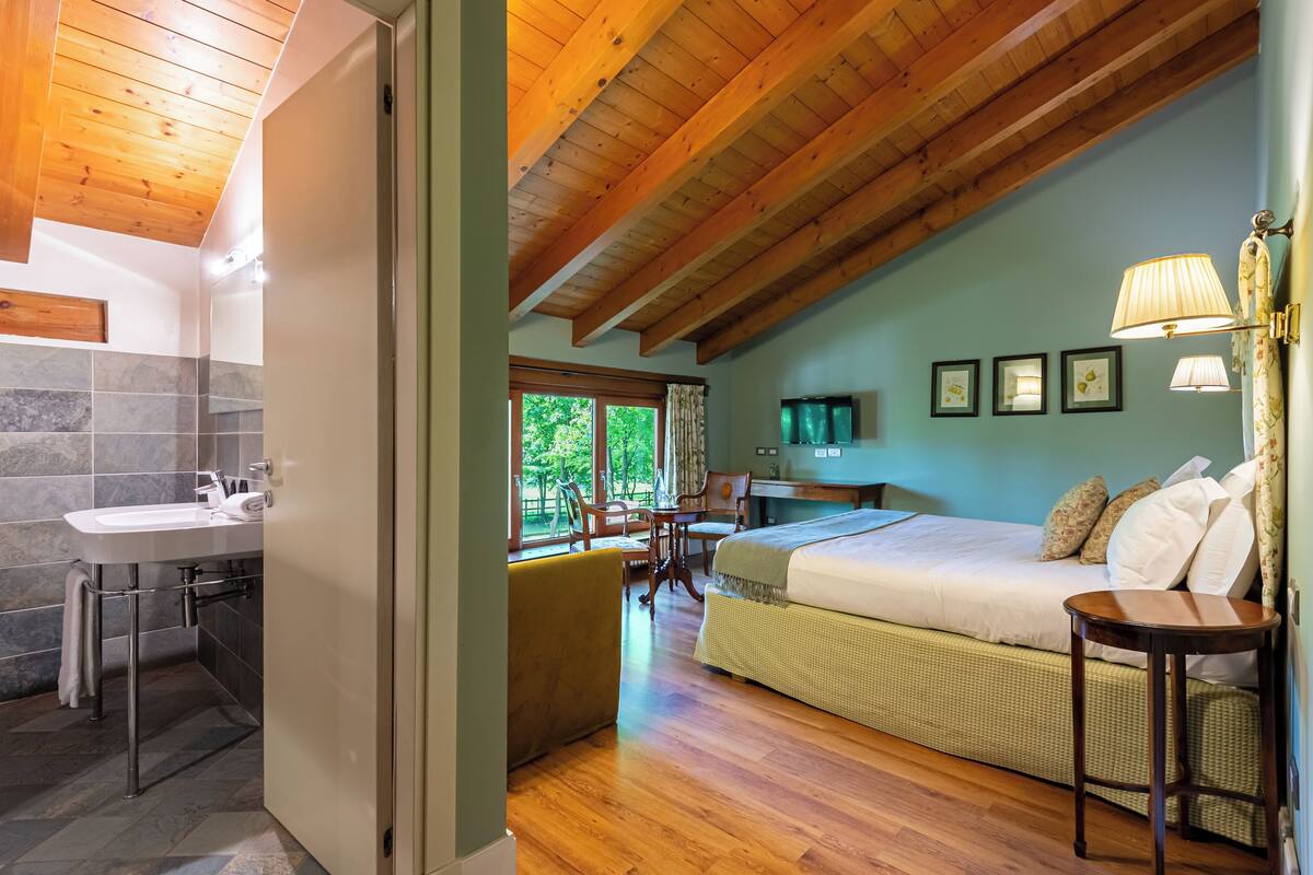 Una habitación con techo de madera, una cama con almohadas, una mesita de noche, una ventana con vistas a la vegetación y un baño con lavabo y azulejos.