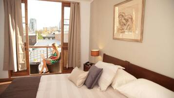 Ein Hotelzimmer mit einem großen Bett, einem Schreibtisch mit einem Laptop, einem Sessel und einem Balkon mit Blick auf die Stadt.