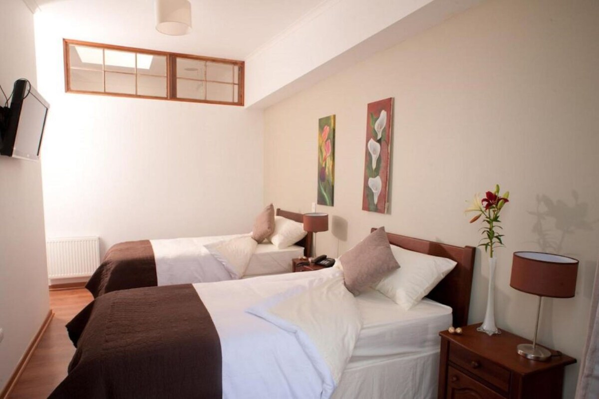 A hotel room with two beds, a window, artwork on the wall, and a nightstand with a lamp.