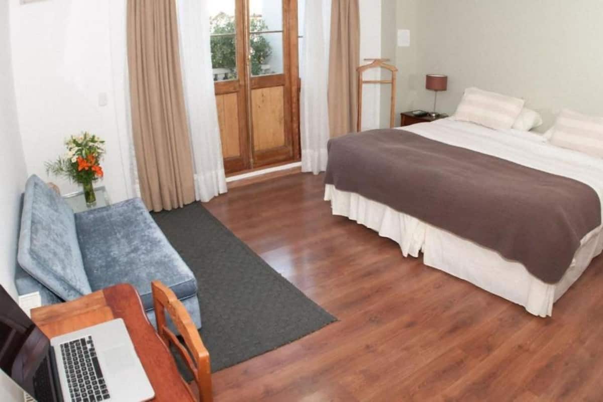A hotel room with a bed, a desk with a laptop, a chair, a wooden floor, and a view through a door.