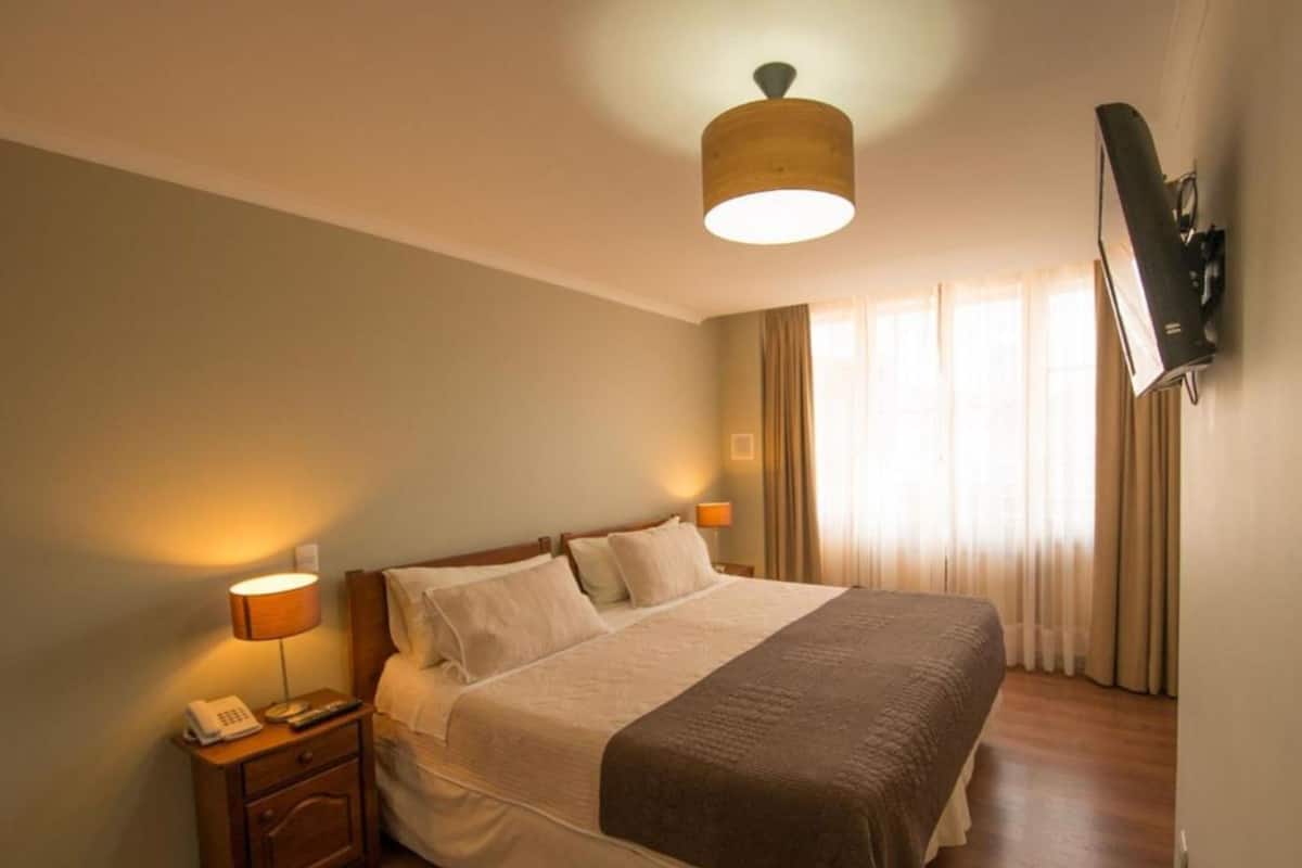 A hotel room with a bed, bedside lamps, a telephone, a television mounted on the wall, and a window with curtains.