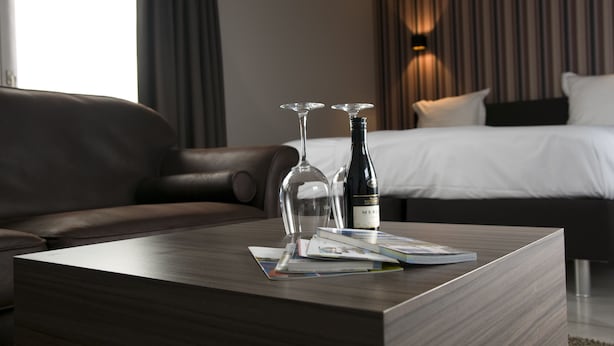 A modern hotel room with a leather sofa, a wooden coffee table with two wine glasses and a bottle, and a neatly made bed with a striped headboard. - Maison Kerkhove (Avelgem)
