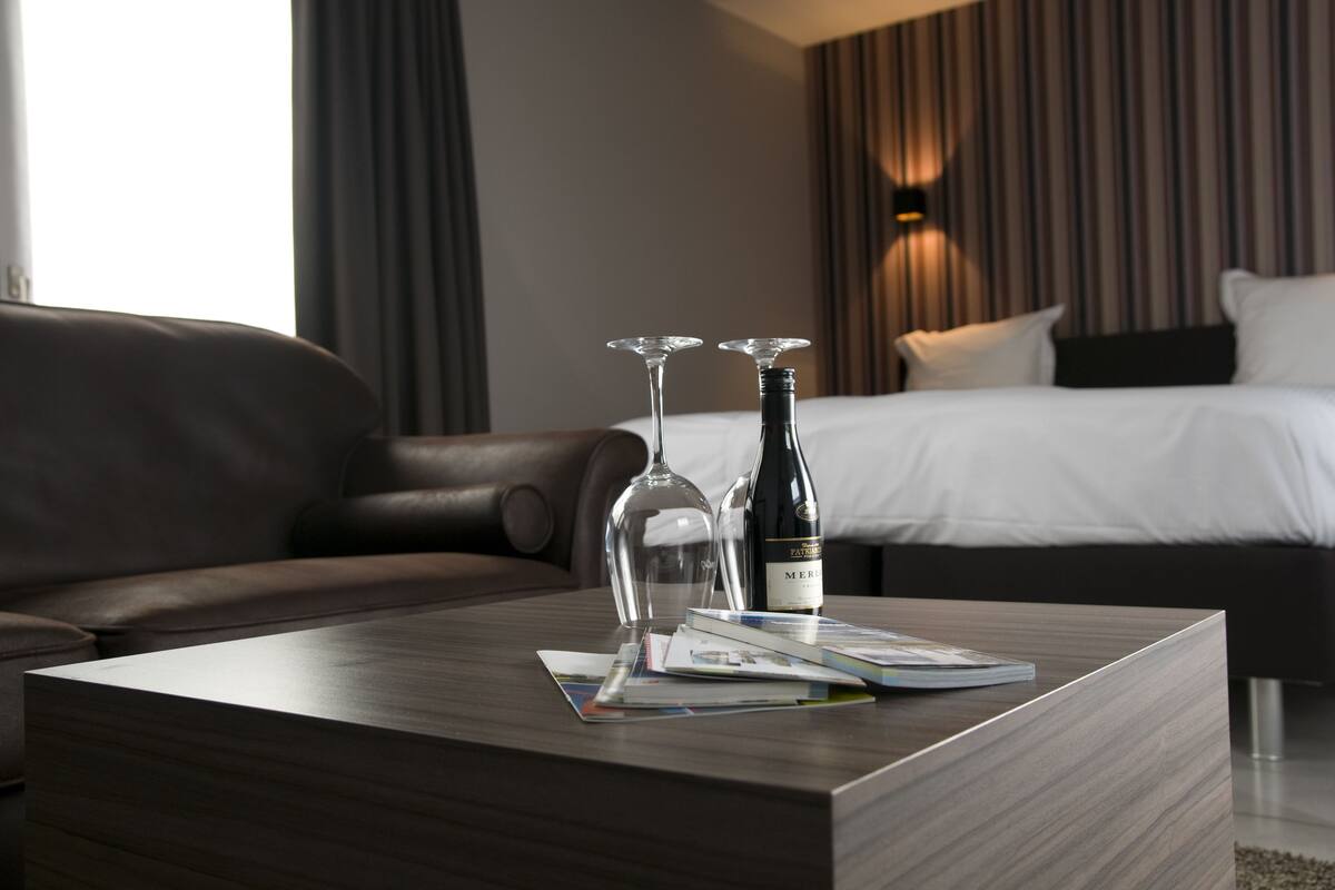 Una habitación de hotel moderna con un sofá de cuero, una mesa de centro de madera con dos copas de vino y una botella, y una cama bien hecha con cabecera a rayas.