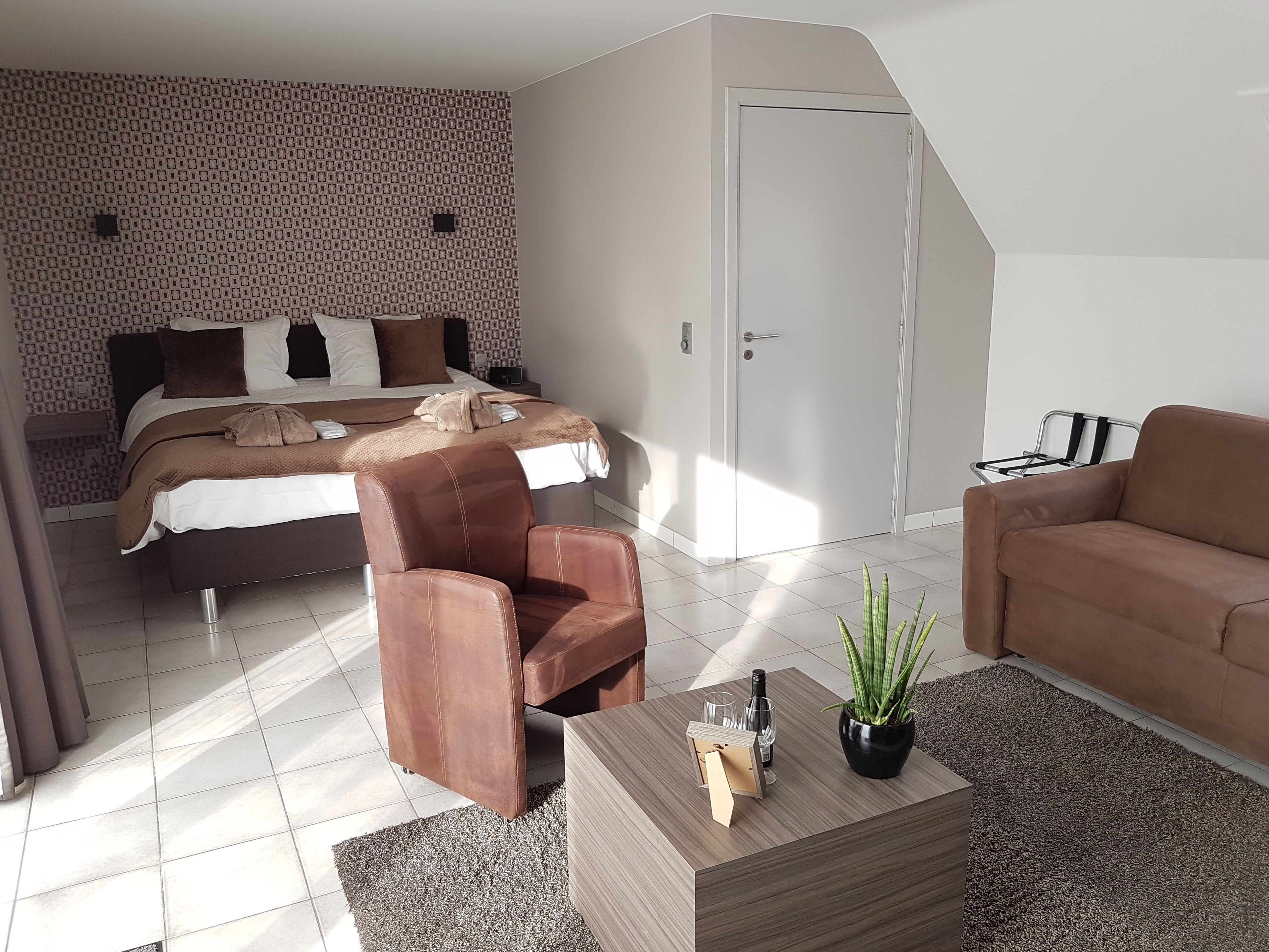 A modern bedroom with a bed, a brown leather chair, a brown sofa, a wooden coffee table with a plant, and a patterned wall.