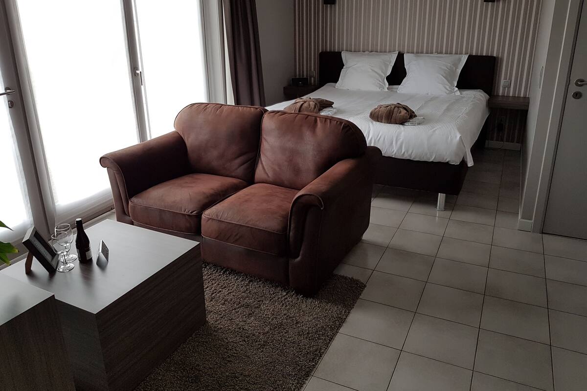 Una habitación de hotel moderna con un sofá de cuero marrón, una pequeña mesa de madera con un vaso y una cama con ropa blanca.