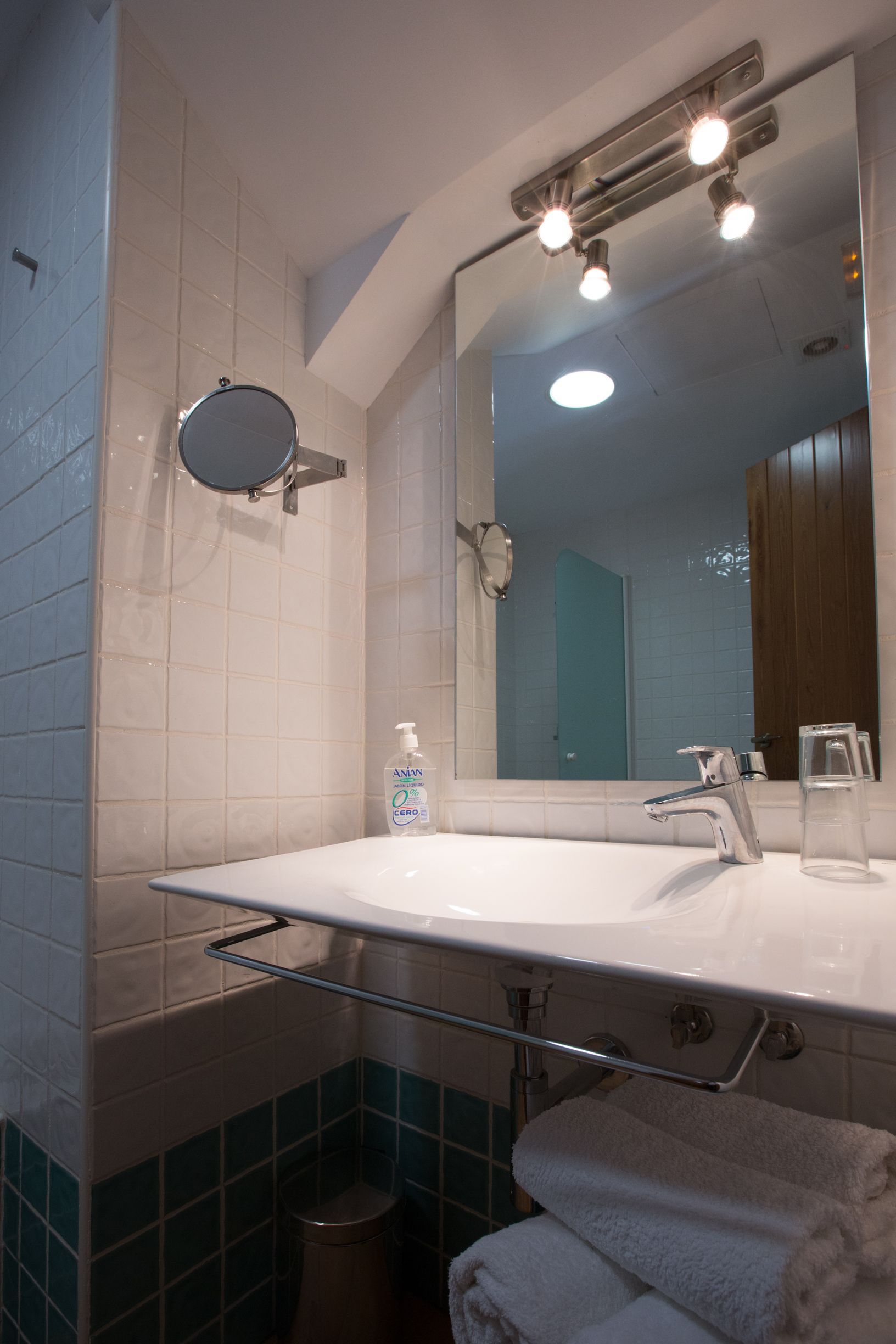 Baño con lavamanos blanco, espejo, espejo de mano, vaso de agua y toallero.