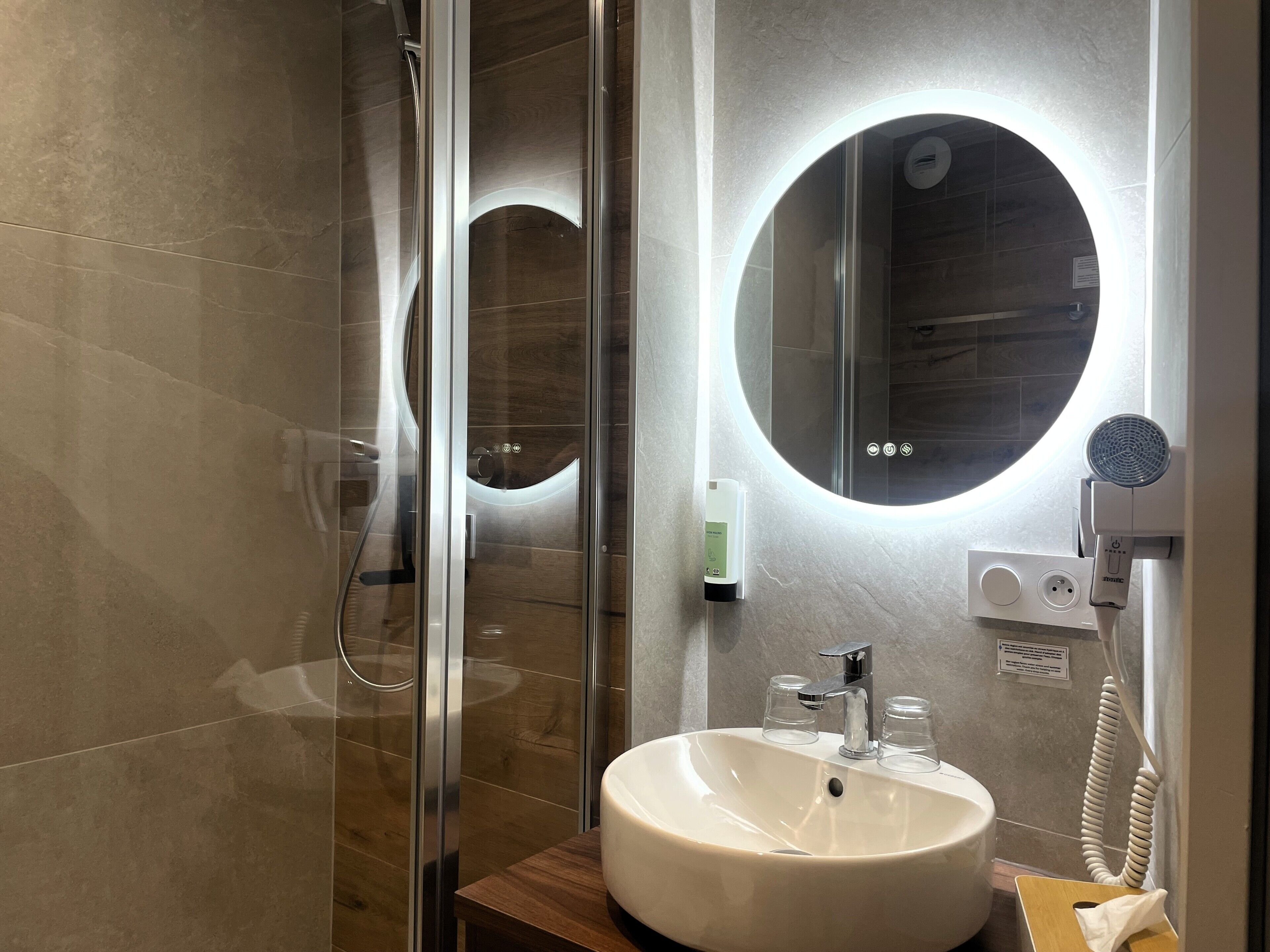 Een moderne badkamer met een douchecabine van glas, een ronde spiegel, een witte wastafel en een handdouche.