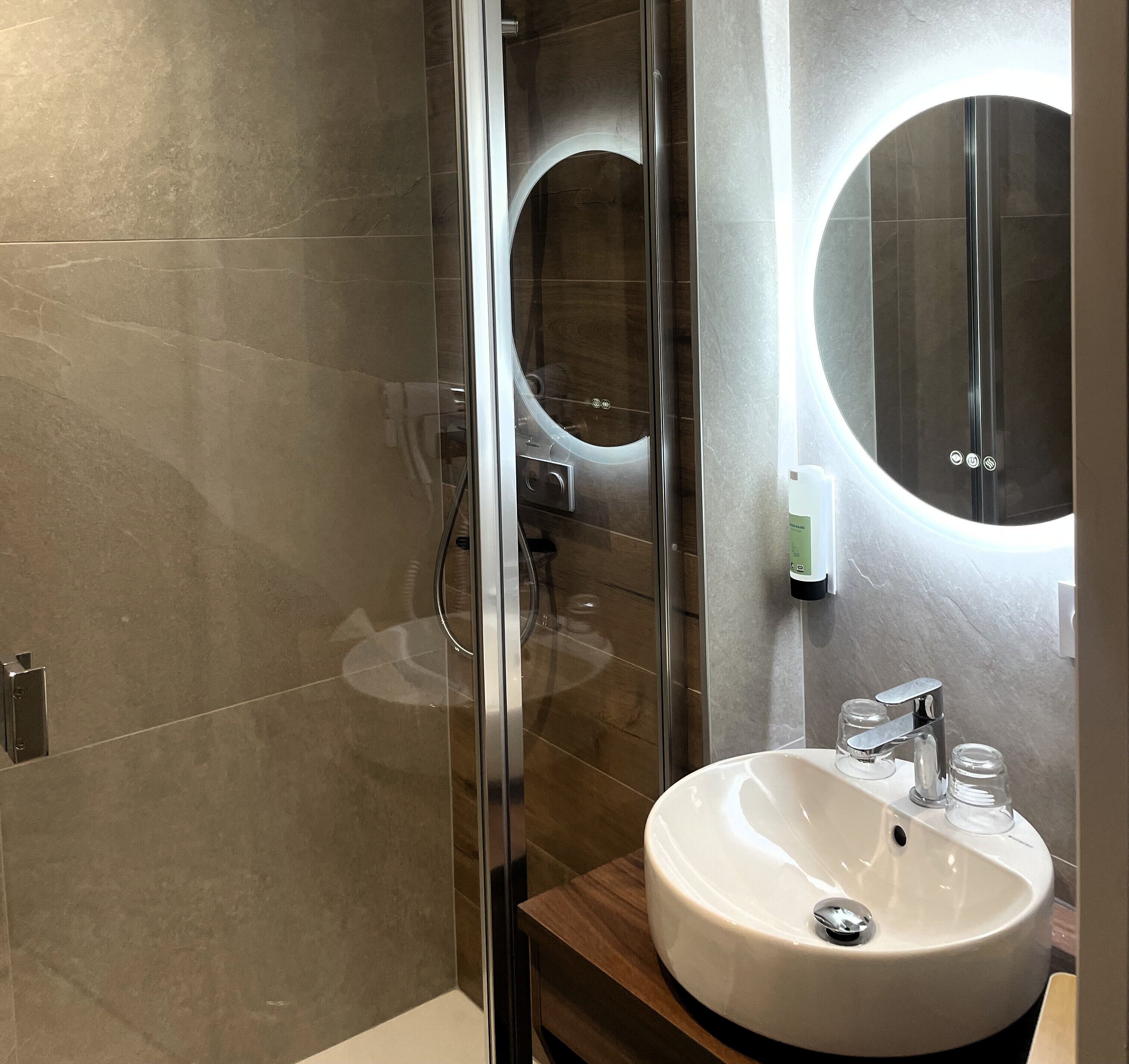 A modern bathroom with a glass shower enclosure, a round mirror, and a white sink with chrome fixtures.