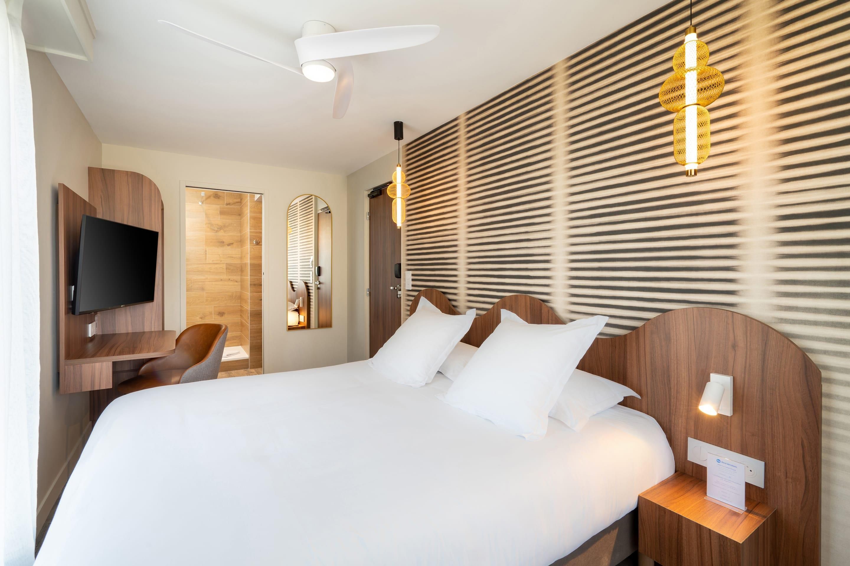 Une chambre d’hôtel avec un grand lit, un téléviseur fixé sur un meuble en bois, un bureau avec une chaise et un luminaire fixé au mur.