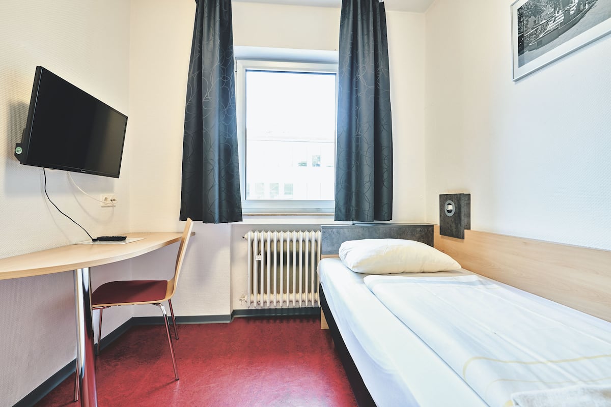 Une chambre d’hôtel comprenant un lit, un bureau avec une chaise, une télévision, une fenêtre avec des rideaux et un radiateur.