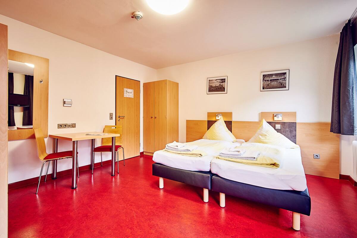 Une chambre d’hôtel avec un sol rouge, un lit avec une literie blanche, une armoire en bois, un bureau avec une chaise et un miroir.
