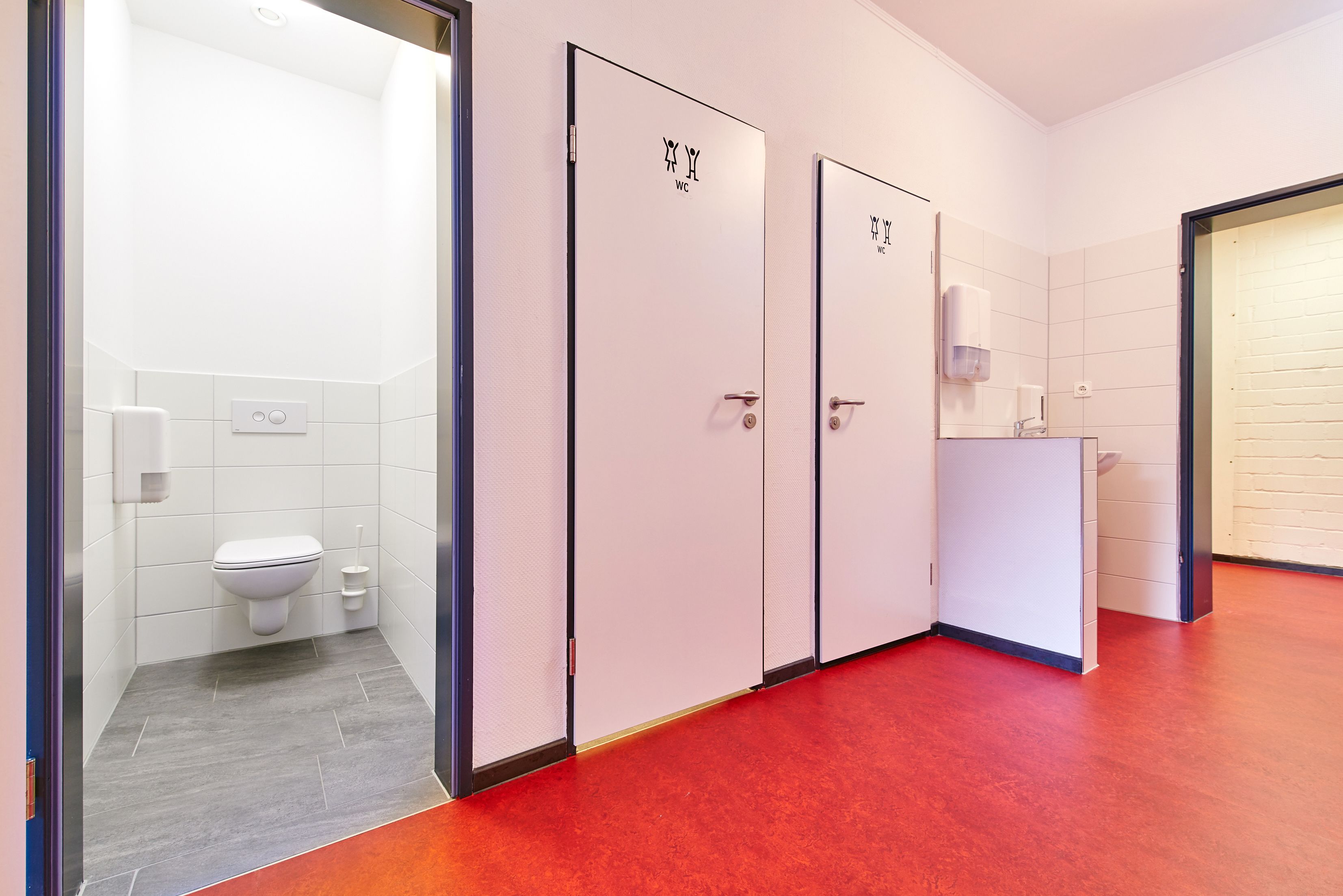 Un pasillo con tres puertas indicadas con la etiqueta 'WC' que dan acceso a un baño con inodoro y secamanos.