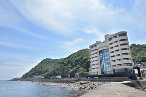 Un edificio costero con fachada curva, costa rocosa y una colina boscosa al fondo.