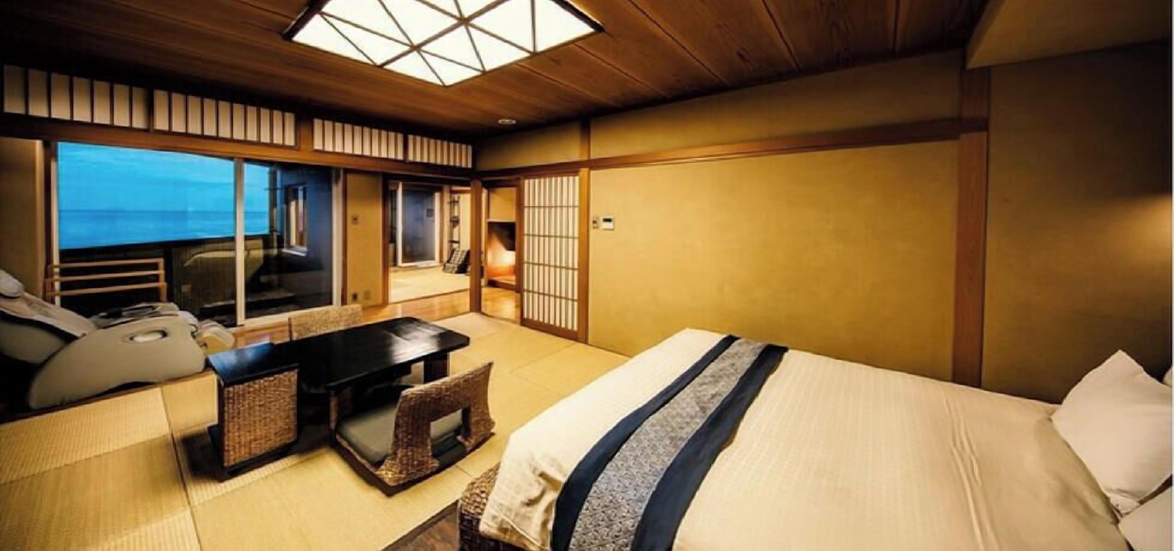 A traditional Japanese-style room with tatami flooring, a bed with a blue and white striped bedspread, a small table with a cushion, and a view of the ocean through large windows.