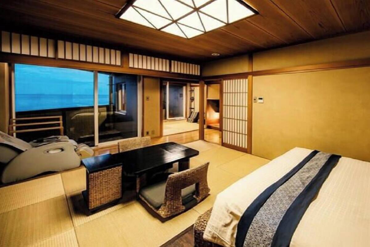 Ein traditioneller japanischer Raum mit Tatami-Boden, einem Bett mit blau-weiß gestreifter Bettdecke, einem kleinen Tisch mit Kissen und Meerblick durch große Fenster.