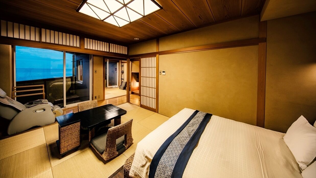 A traditional Japanese-style room with tatami flooring, a bed with a blue and white striped bedspread, a low wooden desk, and a view of the sea through large sliding doors.