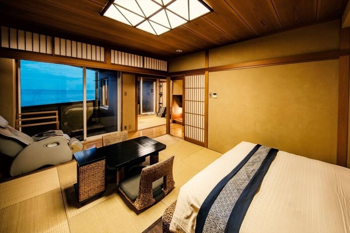 Ein traditioneller japanischer Raum mit Tatami-Böden, einem Bett mit blau-weiß gestreifter Bettdecke, einem niedrigen Holztisch und Meerblick durch große Schiebetüren.