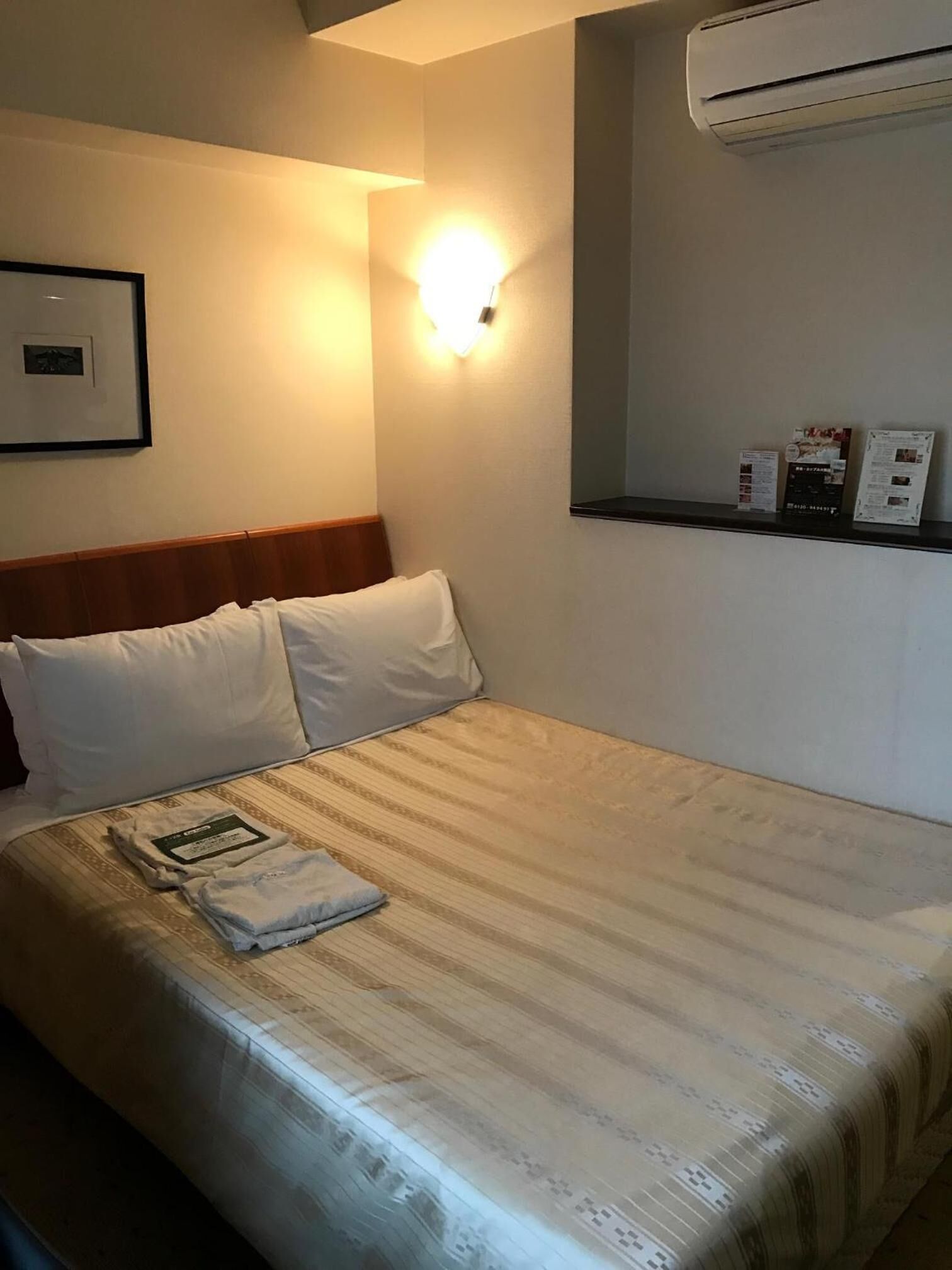 Une chambre d’hôtel équipée d’un lit, d’un climatiseur fixé au mur et d’une étagère contenant des brochures d’information.