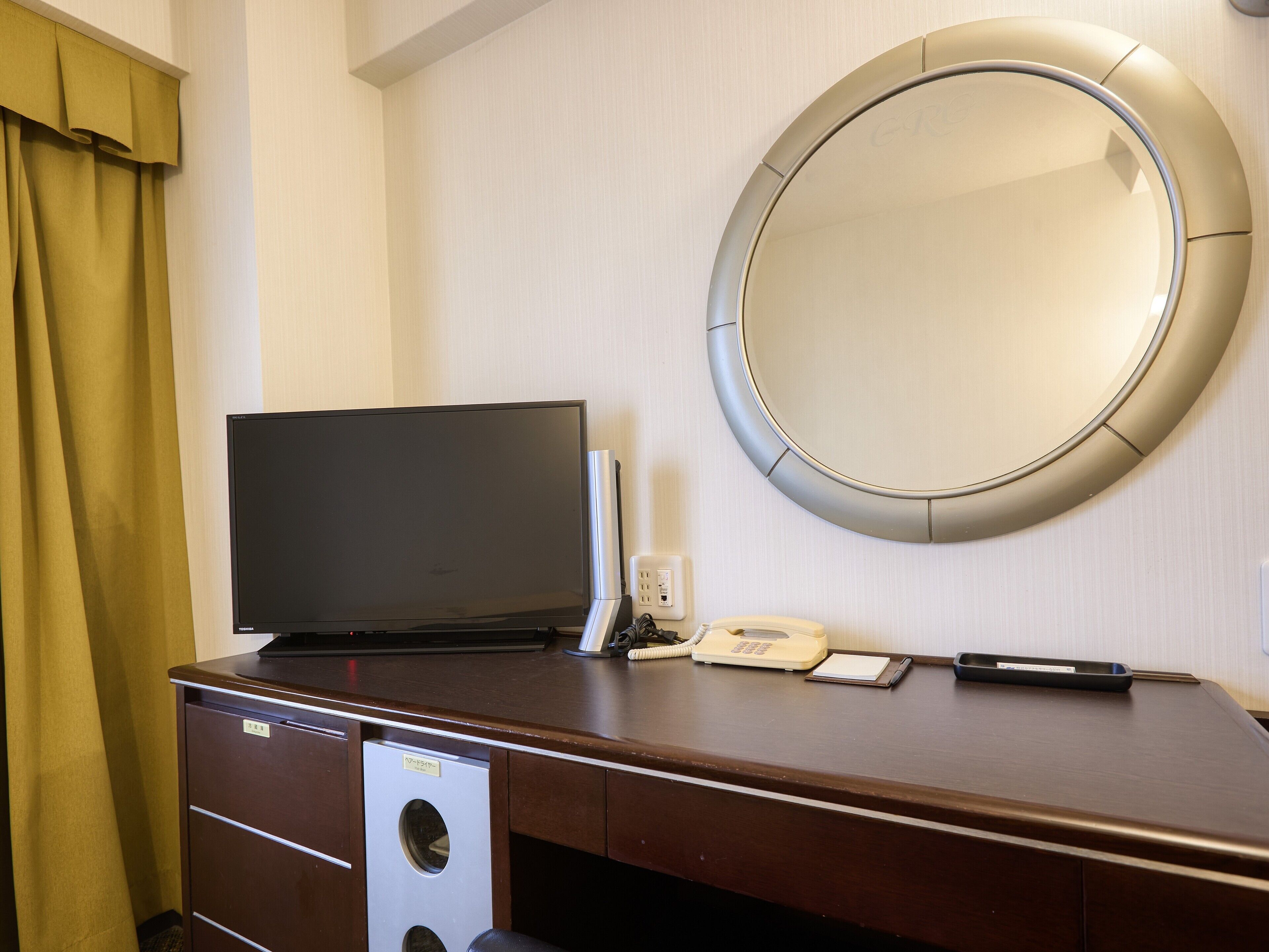A hotel room with a wooden desk, a flat-screen TV, a telephone, and a mirror.
