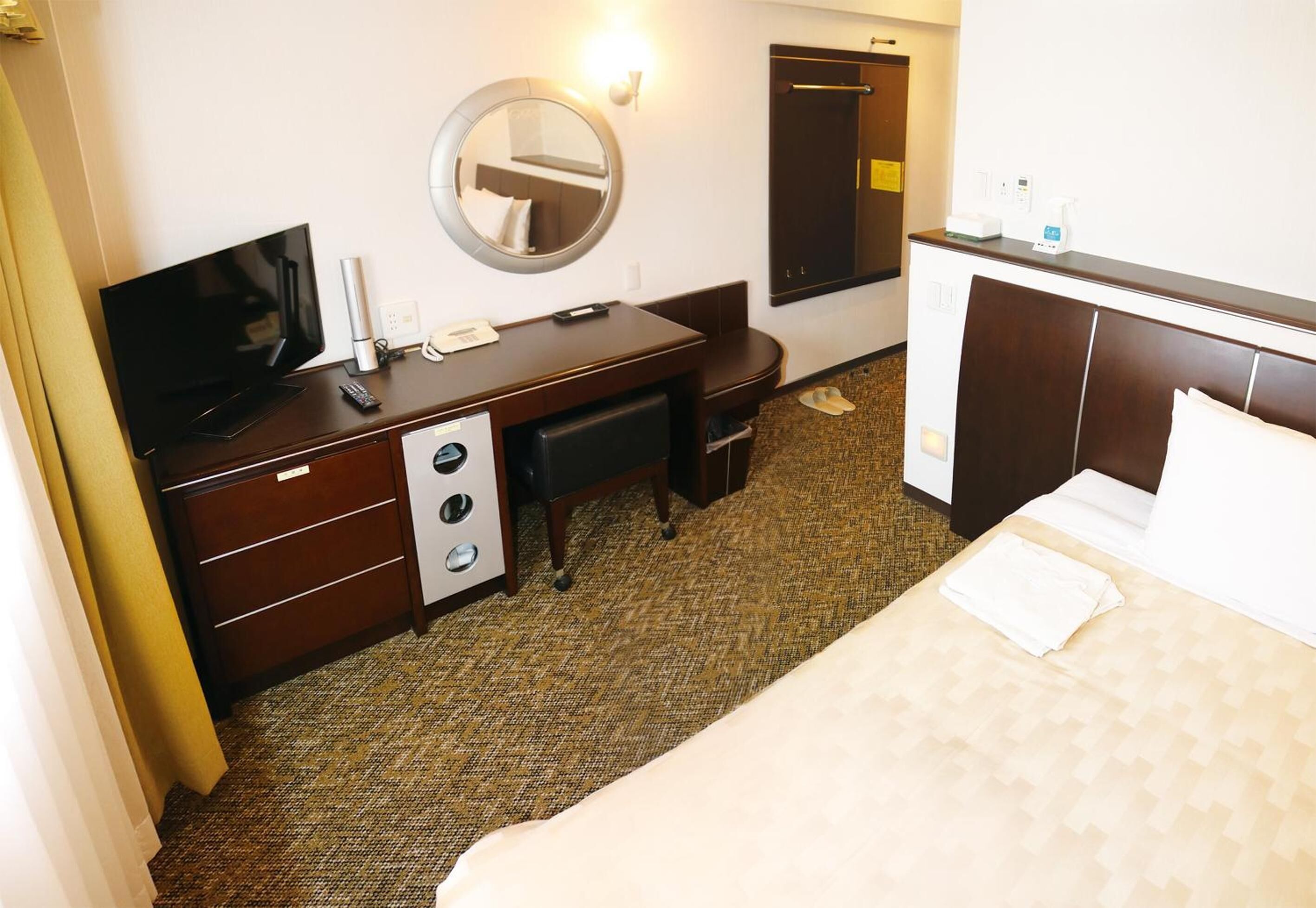 A hotel room with a bed, a desk with a TV, a mirror, a wardrobe, and a bathroom door.