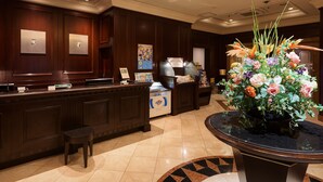 A reception area with a dark wood reception desk, a small stool, a floral arrangement, and a tiled floor. - GRG Hotel Naha (Naha)