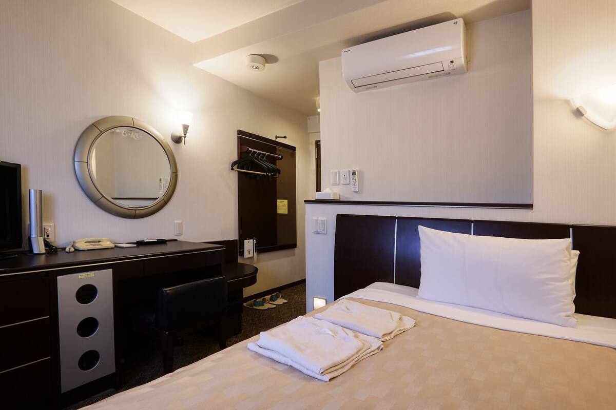 A hotel room with a bed, a desk with a mirror, an air conditioning unit, and a wall-mounted telephone.