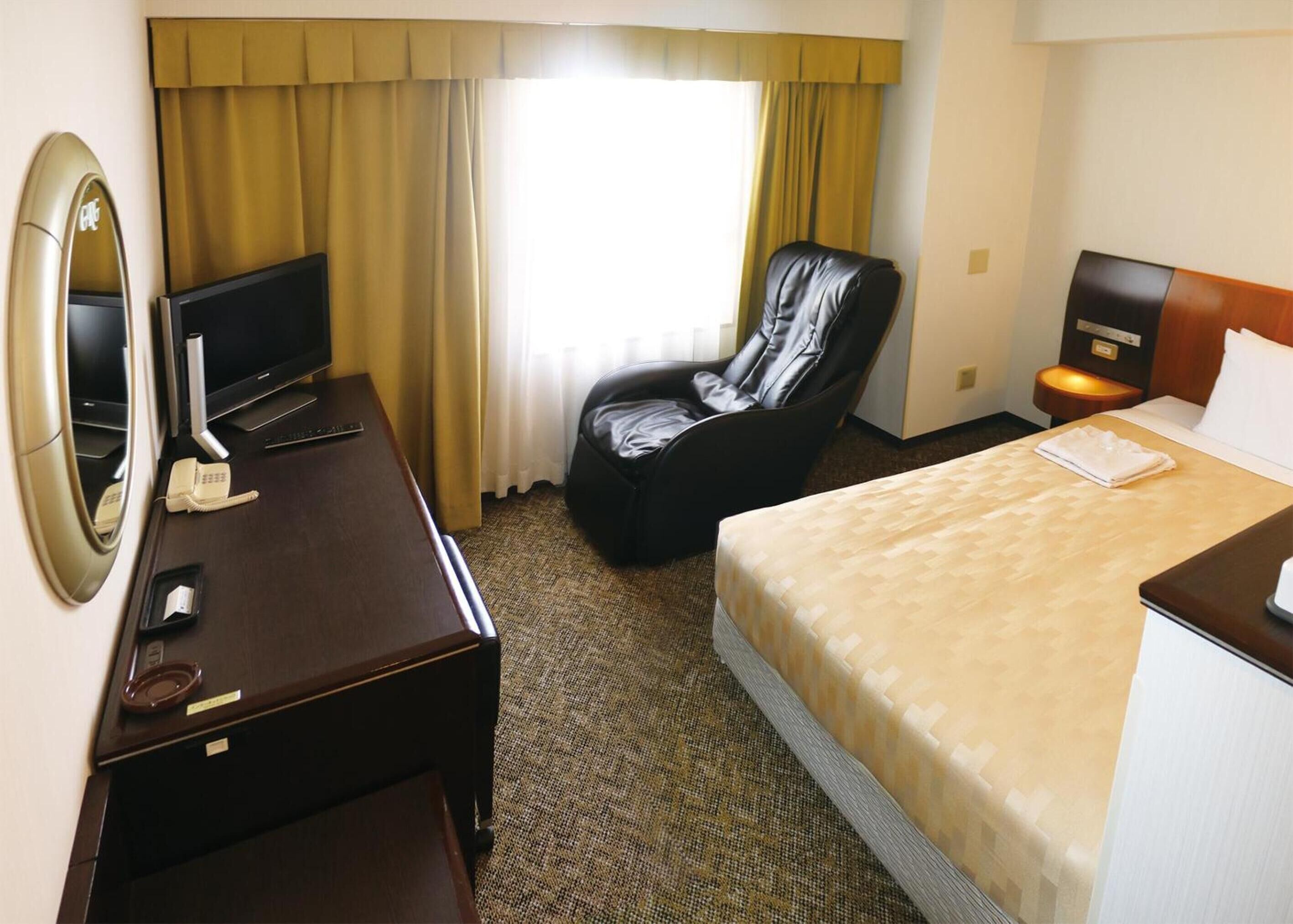 Habitació d'hotel amb un llit, una taula de treball amb un ordinador, una cadira, una tauleta de nit i un mirall.