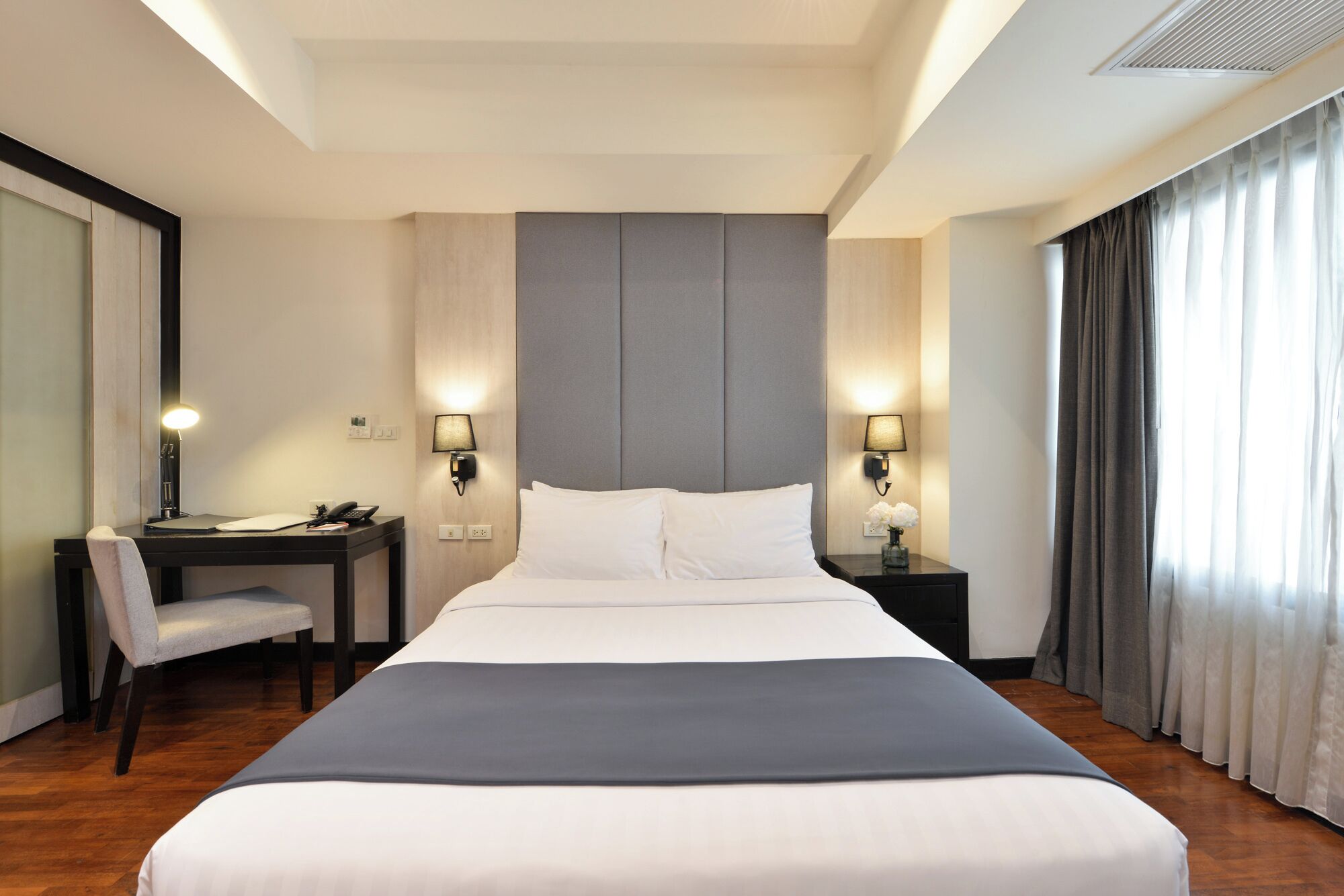 A hotel room with a large bed, a desk, a chair, two bedside lamps, and a window with curtains.