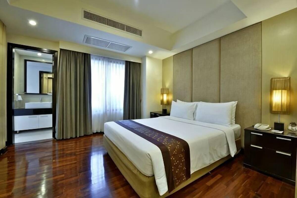 A hotel room with a large bed, a bedside table with a lamp, a nightstand, a mirror, and a bathroom visible through an open door.
