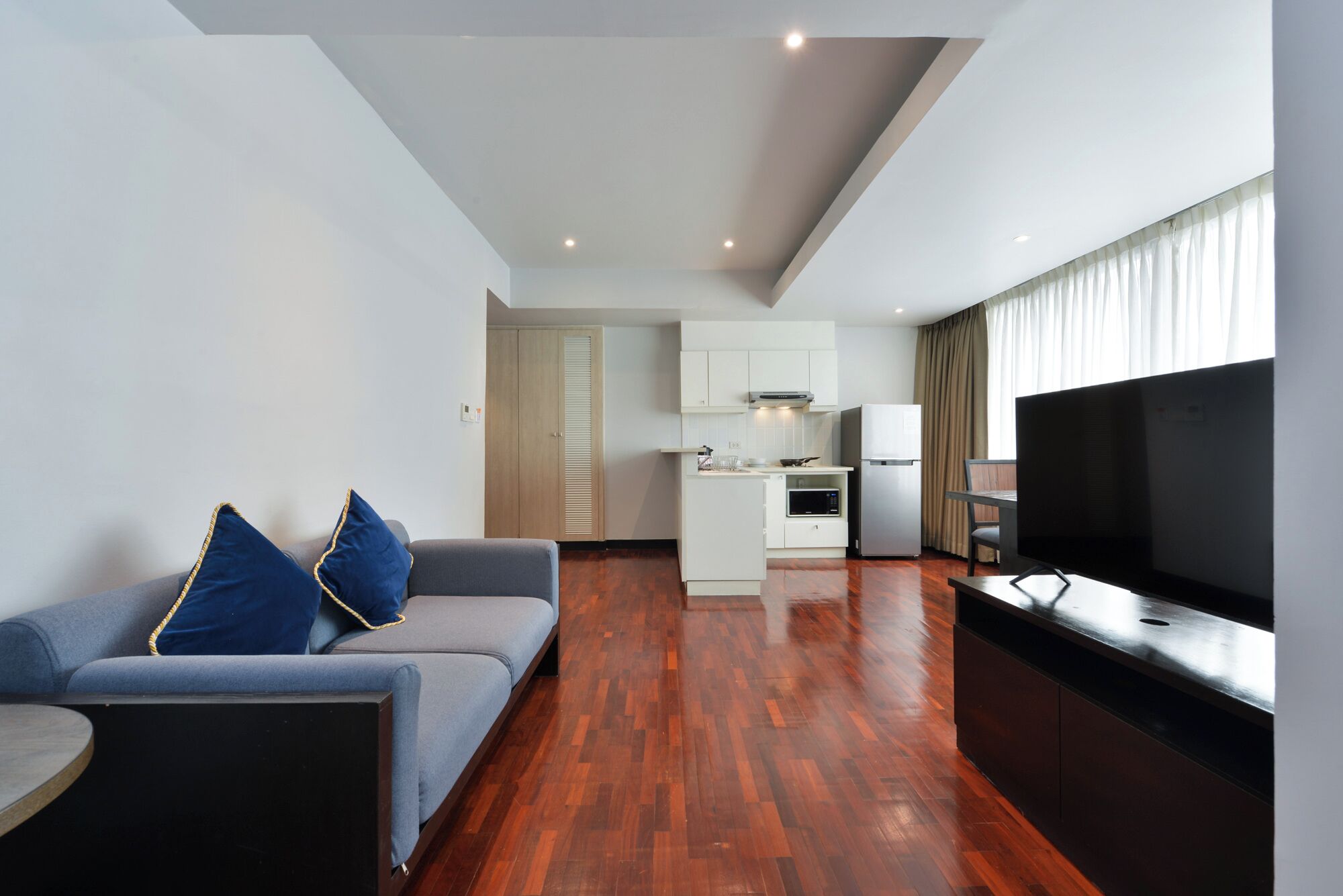 A modern living room with a sofa, a flat-screen TV, and a kitchen area in the background.