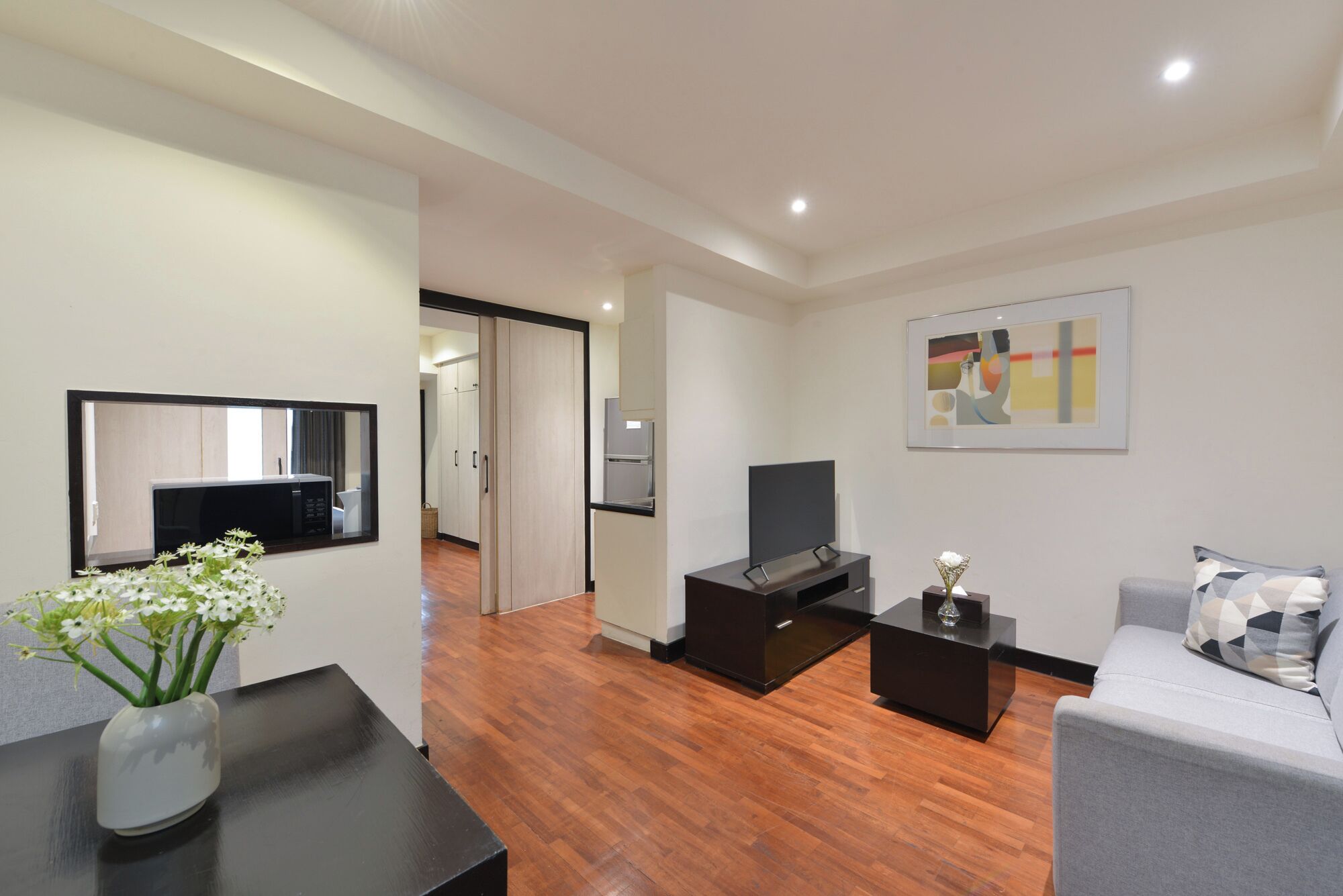 A modern living room with a flat-screen TV, a coffee table, and a sofa. There is a wooden floor and a door leading to a kitchen area.