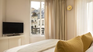 A hotel room with a bed, a television on a stand, a window with curtains, and a view of buildings outside. - Grand Hôtel Richer (Paris)