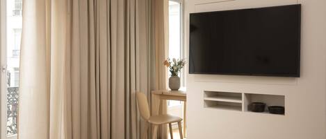 Habitación de hotel con cama, televisor montado en la pared, una silla, una mesa y ventana con cortinas.