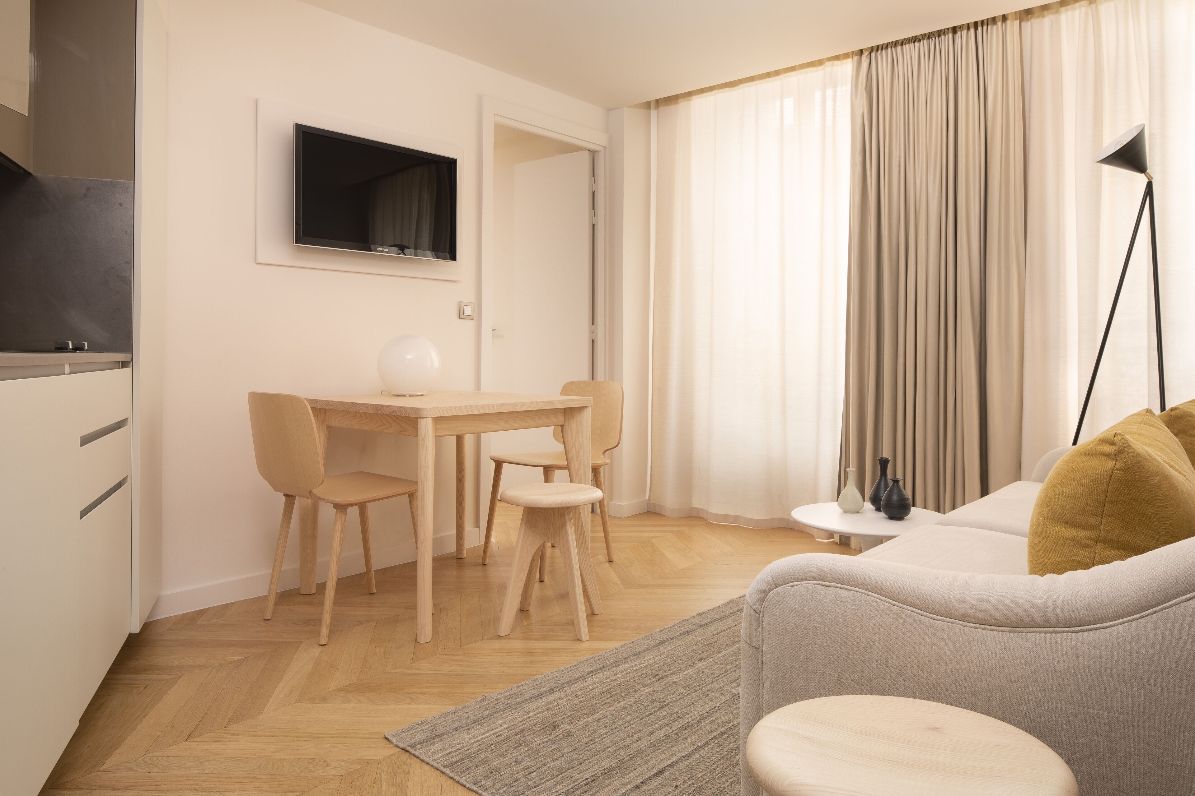 A modern living room with a sofa, a dining table with chairs, a TV mounted on the wall, and a lamp.