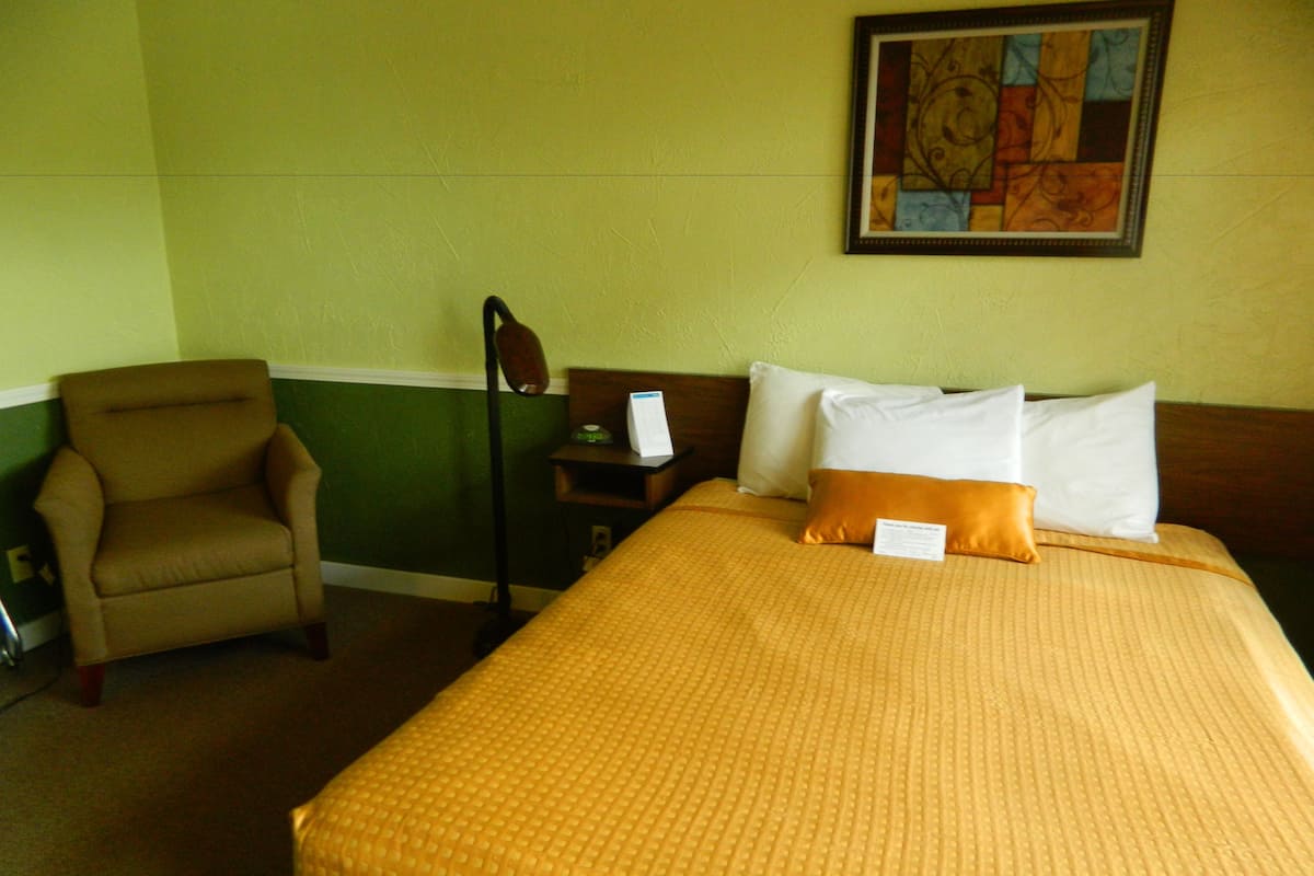 A hotel room with a bed, a chair, a nightstand, and a painting on the wall.