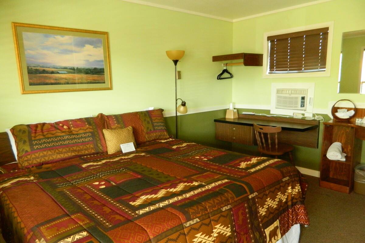 A room with a bed, a desk, a chair, a lamp, a window with blinds, and a painting on the wall.
