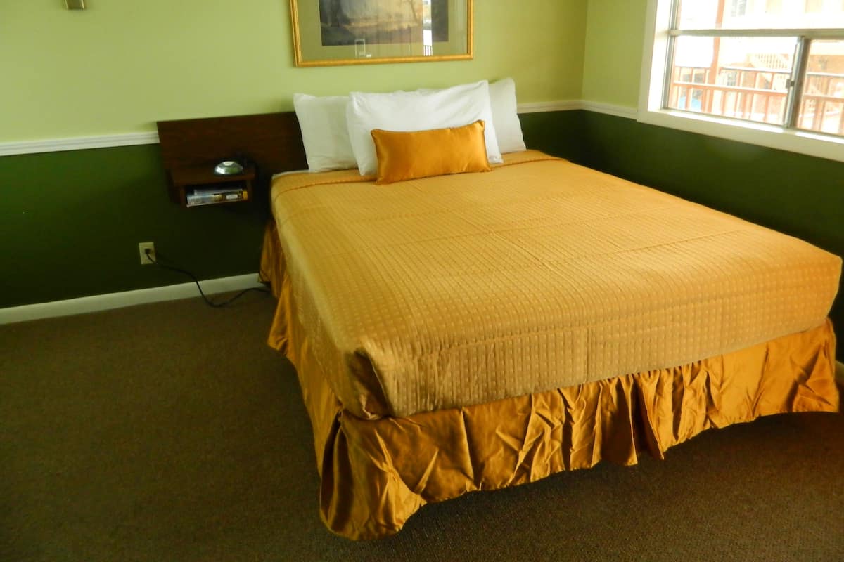 A neatly made bed with a brown bedspread, a white pillow, and a single orange pillow. There is a nightstand with a lamp and a framed picture on the wall above the bed.