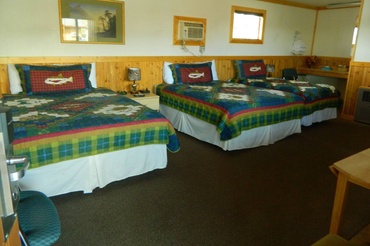 A room with two beds, each with a colorful quilt, a wooden headboard, a small table, and a chair.