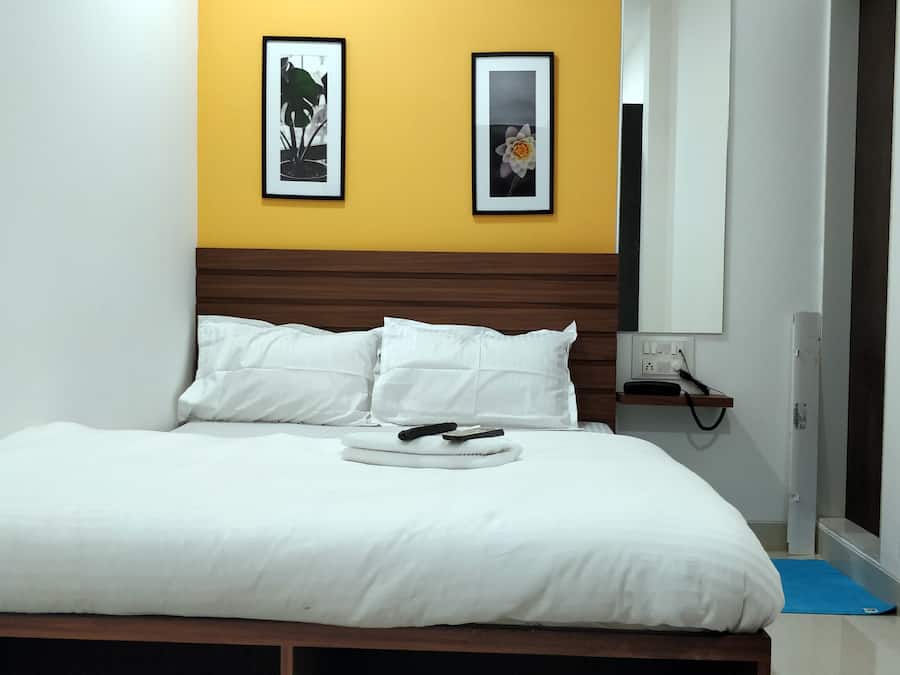 Una cama bien tendida con ropa blanca y almohadas, una mesita de noche con un teléfono y cargador, y dos cuadros en la pared.