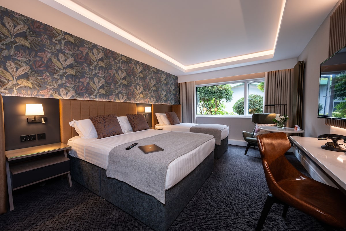 A hotel room with a large bed, a desk, a chair, a small table, and a window with a view of greenery.