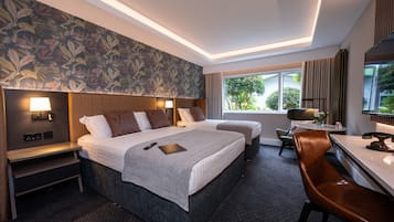 A hotel room with a large bed, a desk, a chair, a small table, and a window with a view of greenery.