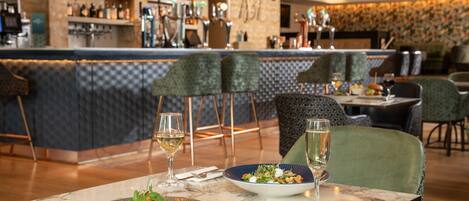 Een restaurant met een bar, marmeren tafels en groene beklede stoelen.
