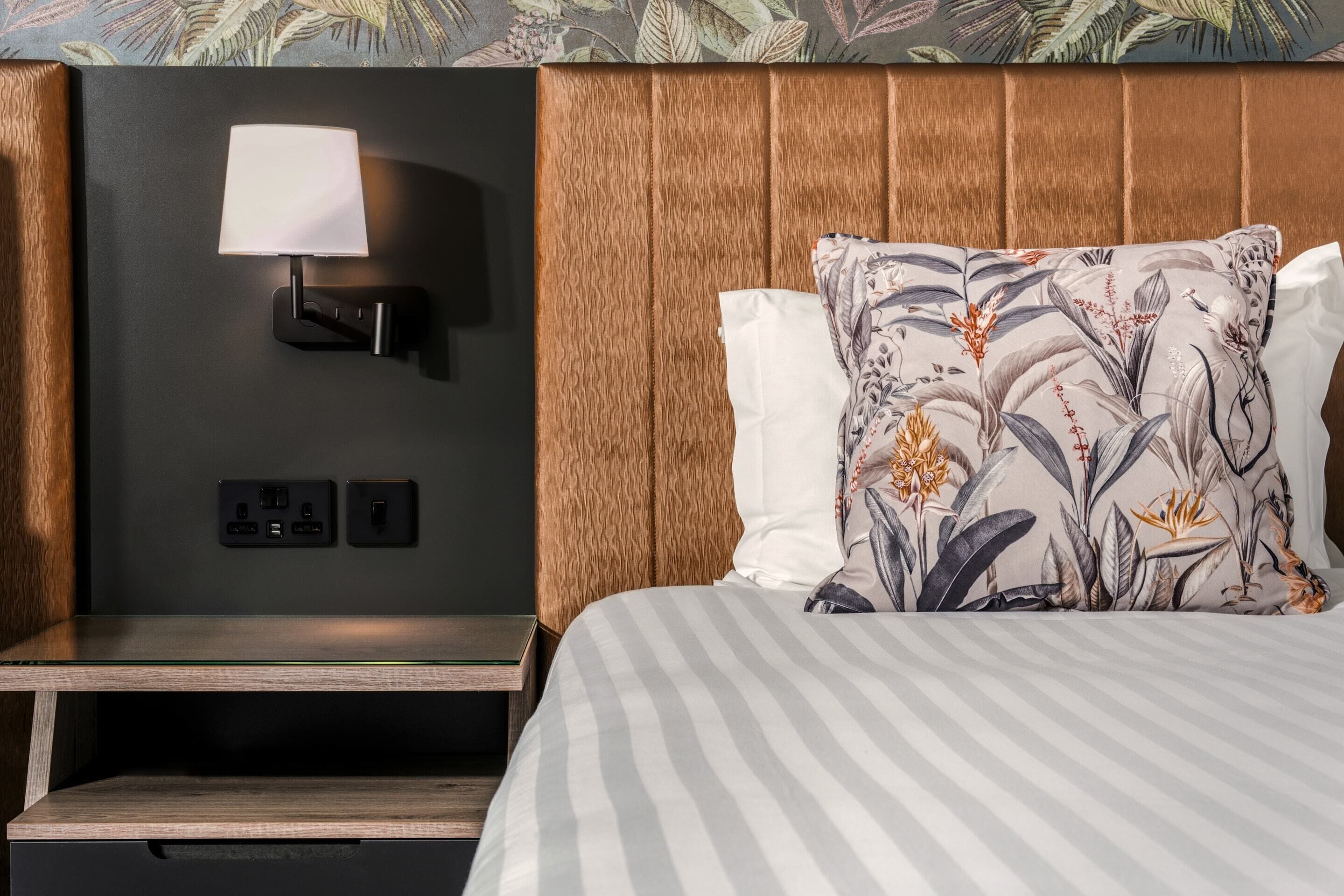 Quarto de hotel com cama, mesa de cabeceira, luminária na parede e tomada.