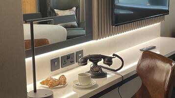 Habitación de hotel de diseño moderno, con una amplia televisión de pantalla plana en la pared, un escritorio con teléfono, una silla y una pequeña mesa con un croissant y una taza.