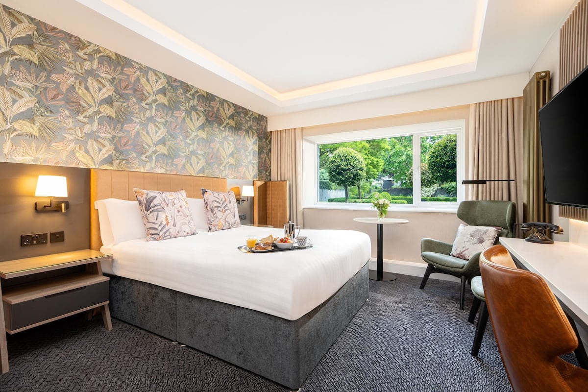 A hotel room with a large bed, bedside tables, a desk, and a chair. There is a window with a view of greenery outside.