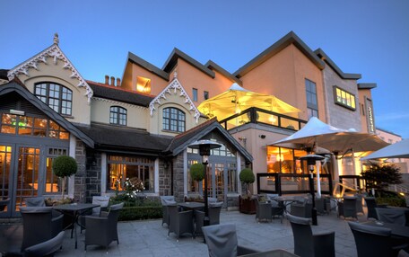 Un patio al aire libre con muebles de mimbre y sombrillas, un edificio de piedra con estilo arquitectónico tradicional, y un cielo vespertino despejado.