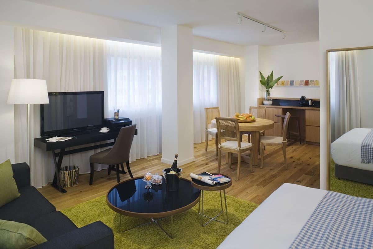A modern hotel room with a flat-screen TV, a dining area, a bed, and a small kitchenette.
