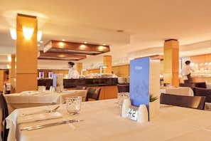 A restaurant setting with tables set for dining, a menu stand, and a chef in the background. - Zafiro Cala Mesquida (Capdepera)
