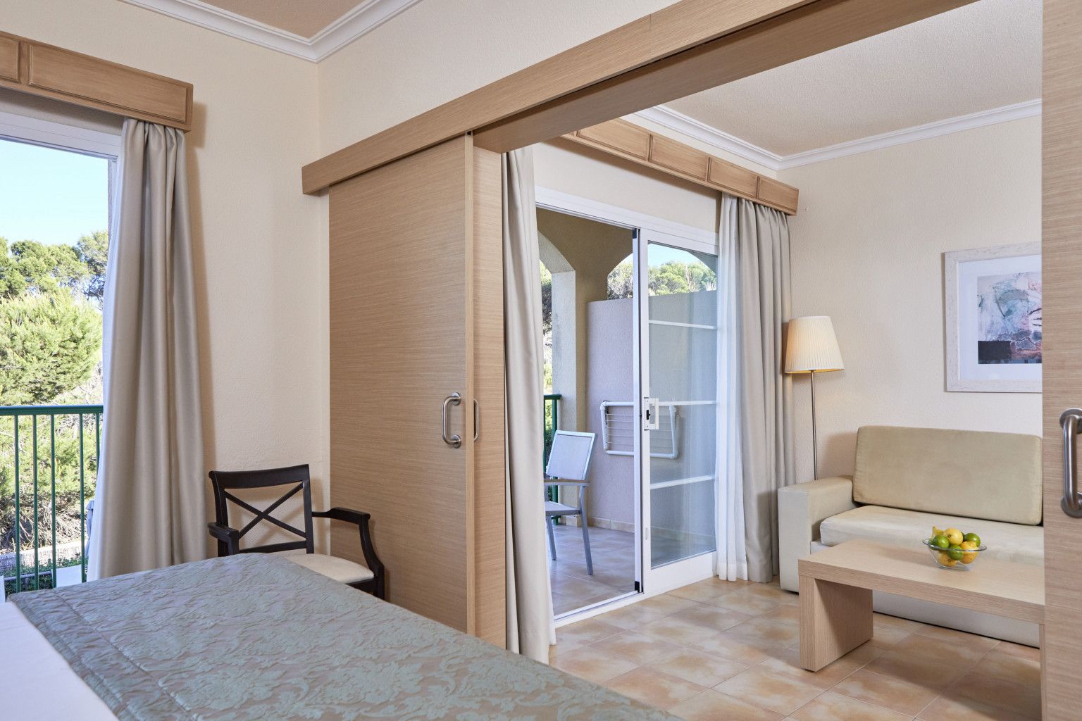 A hotel room with a bed, a sofa, a small table with a fruit bowl, a chair, a sliding door to a balcony, and a lamp.